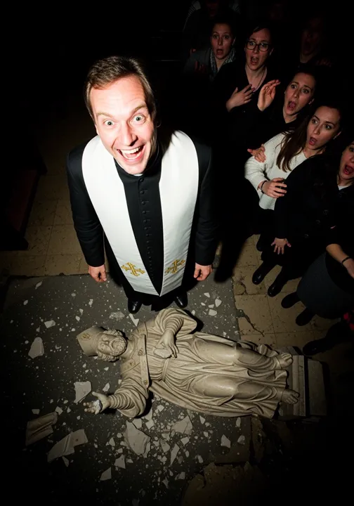 Un cura sonríe desencajado junto a la estatua de un santo hecha añicos en el suelo, ante feligreses espantados.